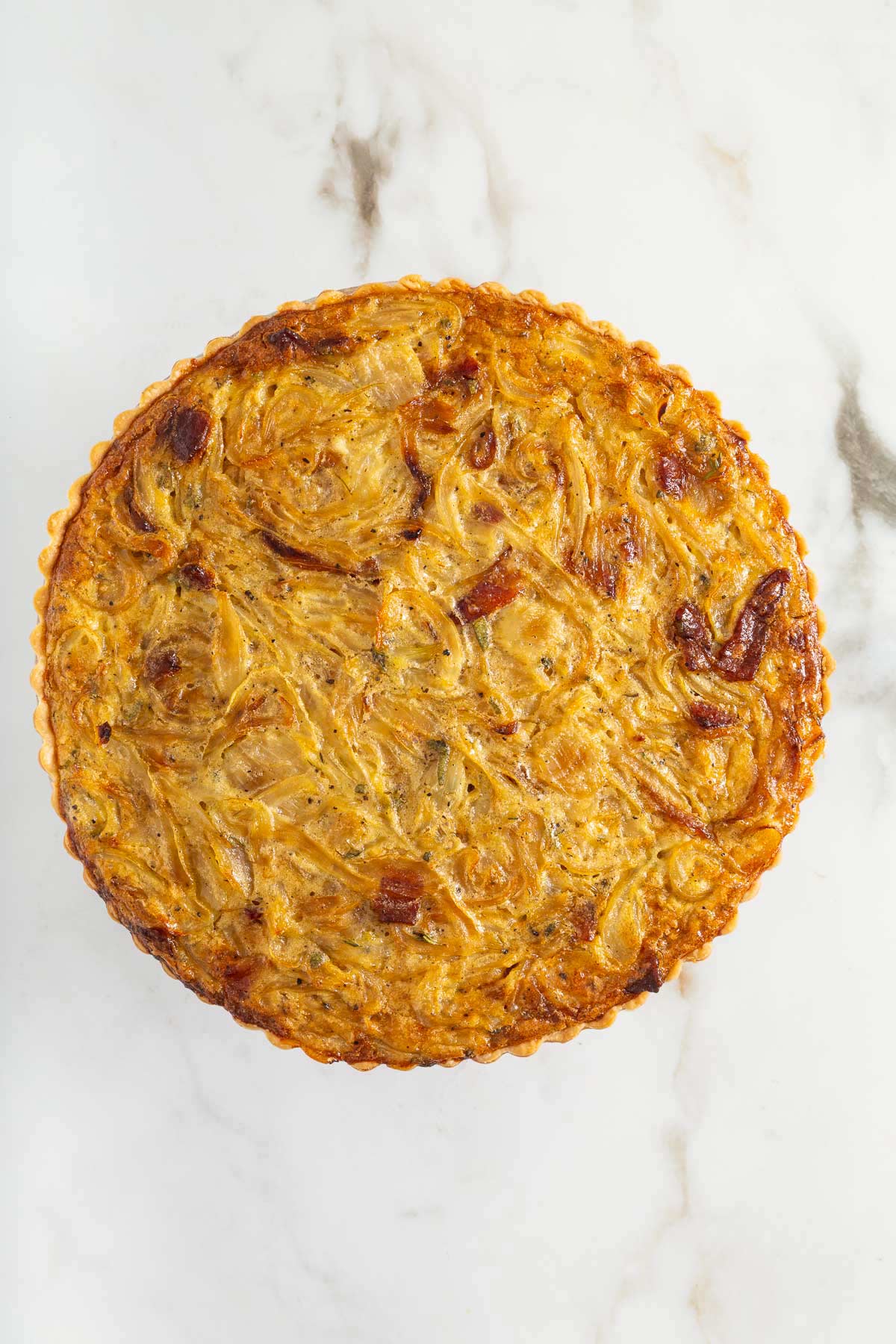 A round, golden-brown onion tart with a flaky crust sits on a white marble surface, inviting you to enjoy the same effortless elegance as an easy sheetpan pancakes recipe.