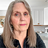 Woman with long gray hair wearing a black shirt, standing in a bright kitchen with white cabinets in the background.
