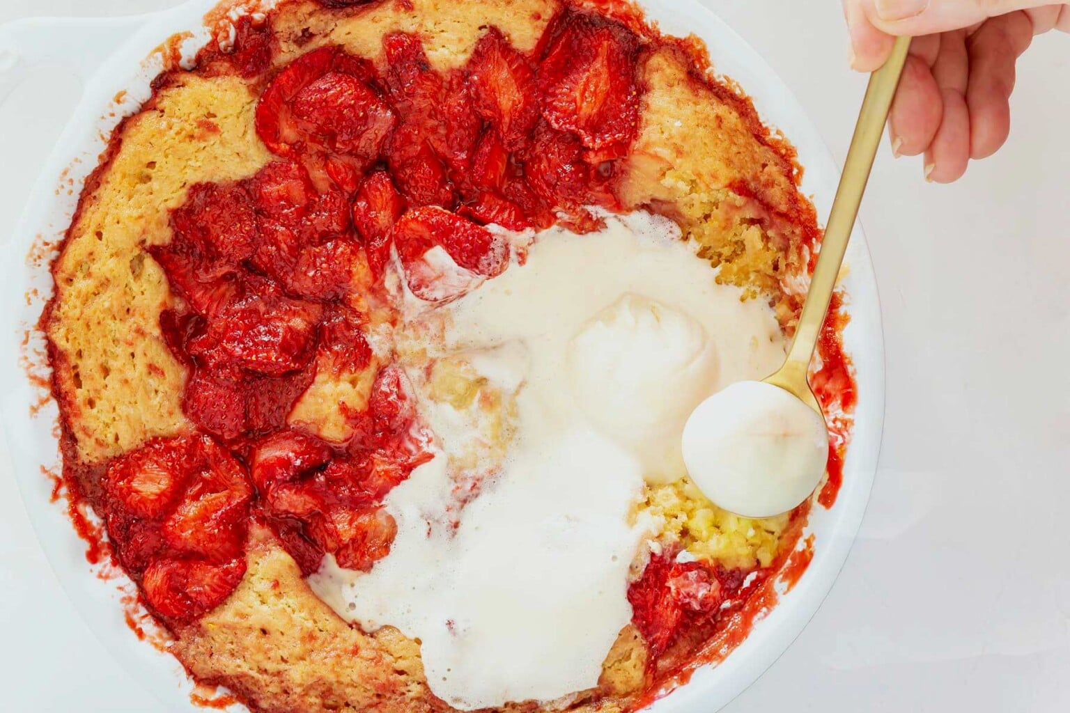 A hand scoops vanilla ice cream onto strawberry cobbler in a white baking dish, with baked strawberries and golden crust visible—reminiscent of an easy strawberry spoon cake, where sweet berry filling meets warm, buttery layers for the perfect dessert.