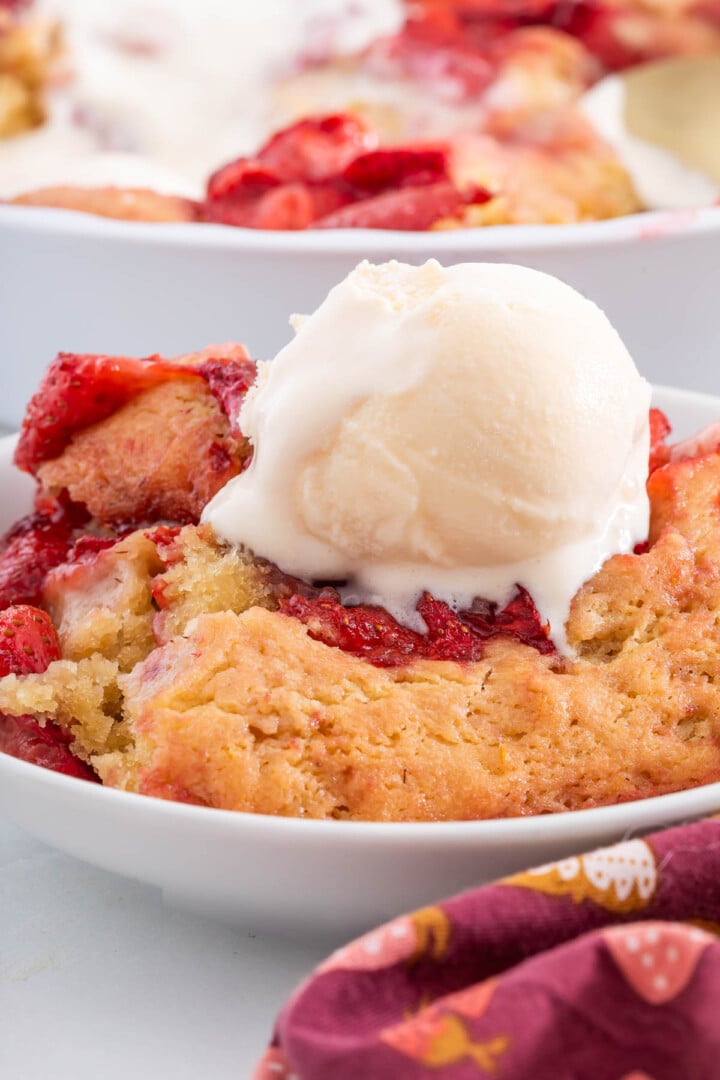 A bowl of strawberry cobbler topped with a scoop of vanilla ice cream, with part of a patterned napkin visible in the foreground.