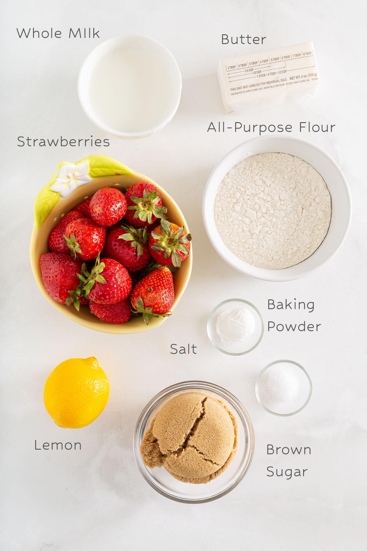 Top-down view of whole milk, butter, all-purpose flour, strawberries, lemon, baking powder, salt, and brown sugar in separate containers on a white surface-perfect ingredients for making easy sheetpan pancakes.