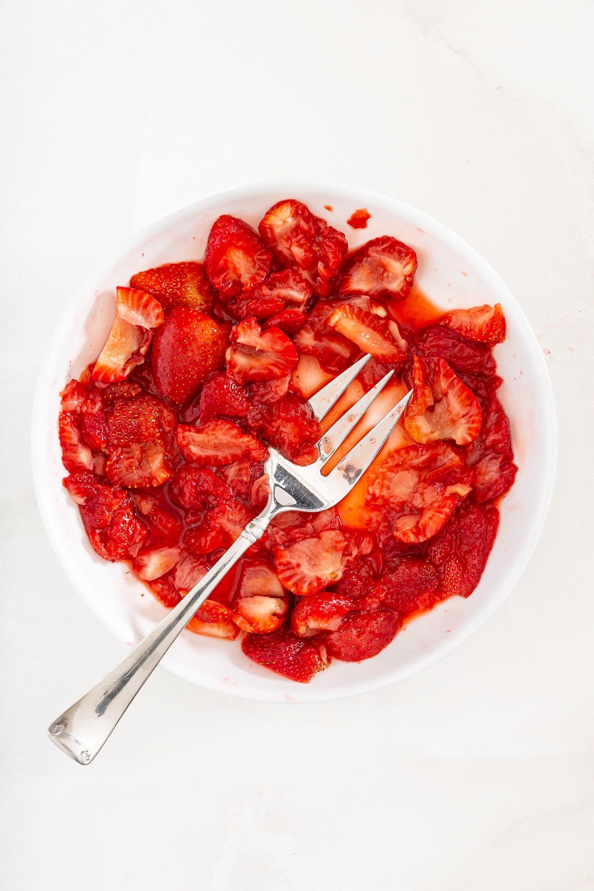 Bowl of sliced and mashed strawberries with a fork, on a white surface-perfect for topping your favorite sheetpan pancake recipe.