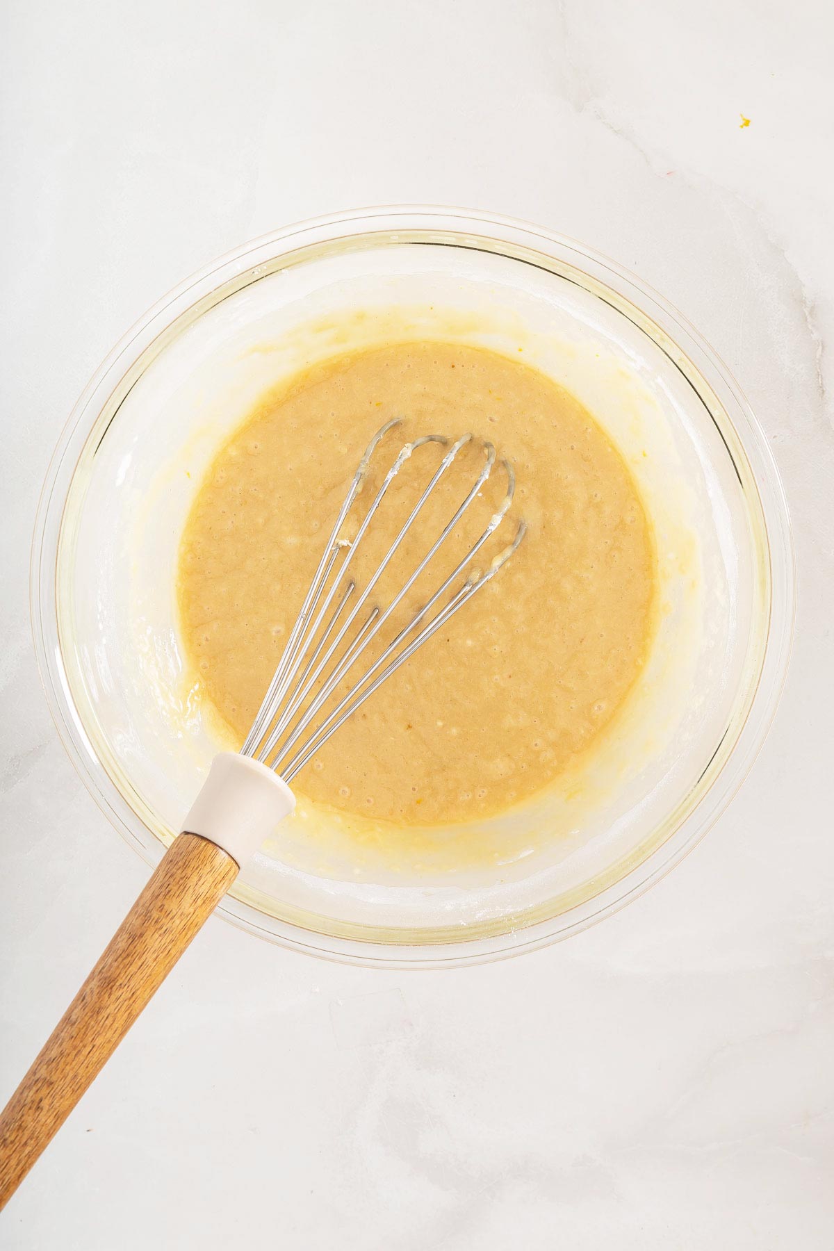 A glass bowl containing yellow batter, perfect for making easy sheetpan pancakes, is shown with a whisk resting inside on a light countertop.