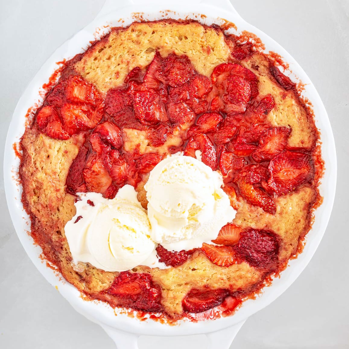 Baked strawberry cobbler topped with two scoops of vanilla ice cream in a white dish, photographed from above-a delicious treat that shares the comforting, crowd-pleasing appeal of classic sheet pan pancakes.