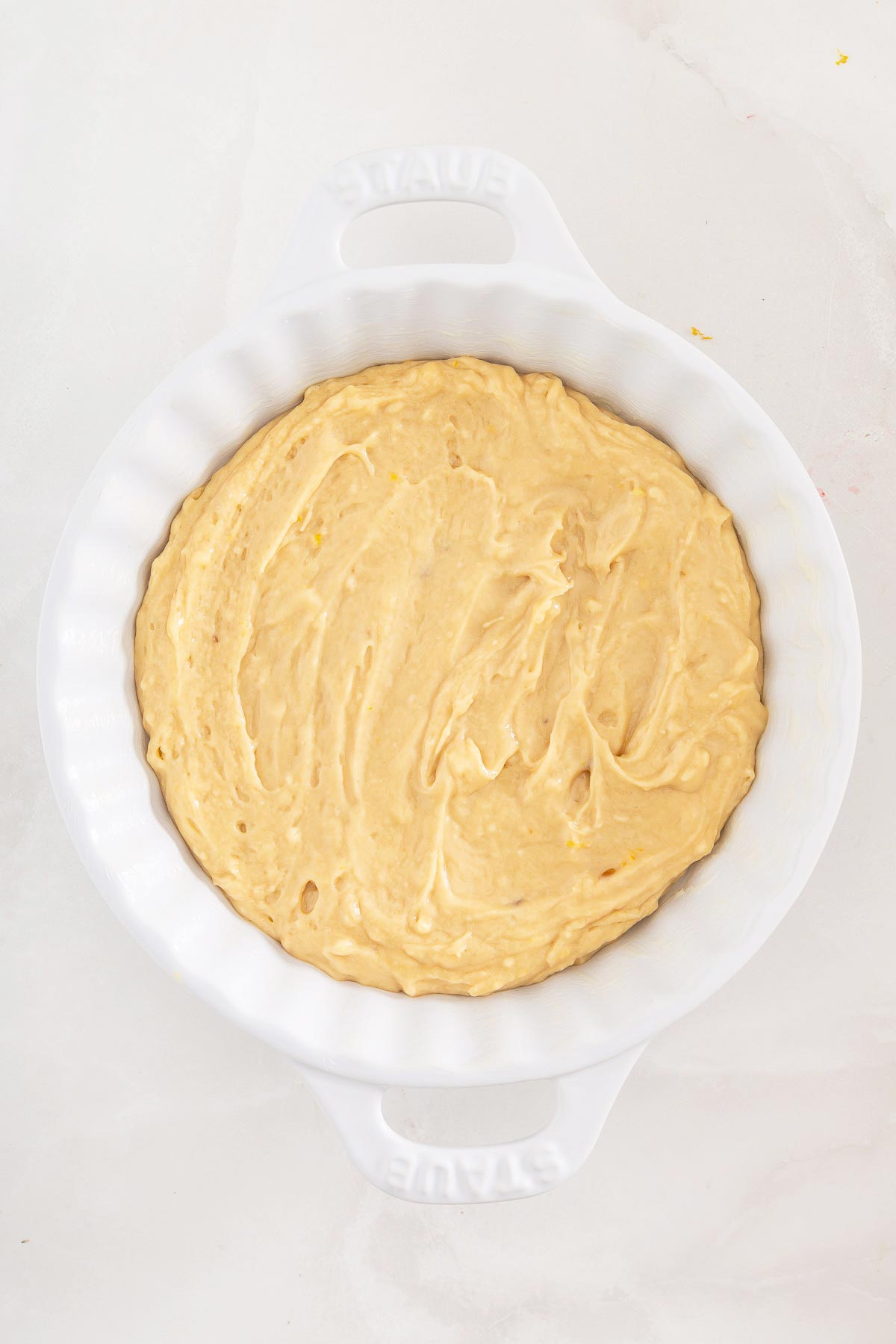 A white round baking dish filled with an even layer of unbaked yellow cake batter, viewed from above on a light surface-perfect for making easy sheetpan pancakes from mix.