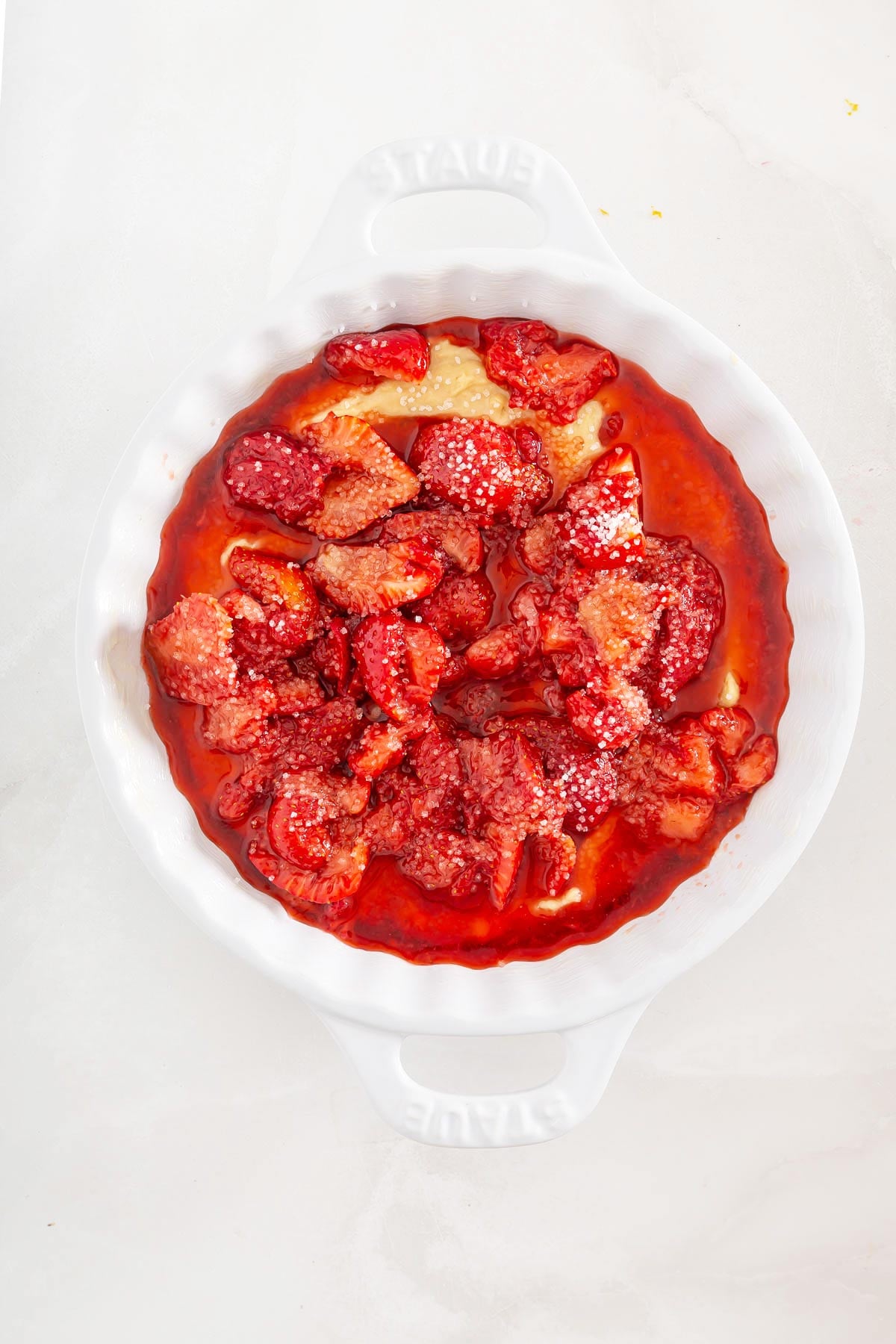 A white baking dish filled with baked strawberries and a golden pastry base, lightly dusted with powdered sugar, on a light-colored surface-a delightful twist reminiscent of easy sheetpan pancakes, perfect for your next brunch.