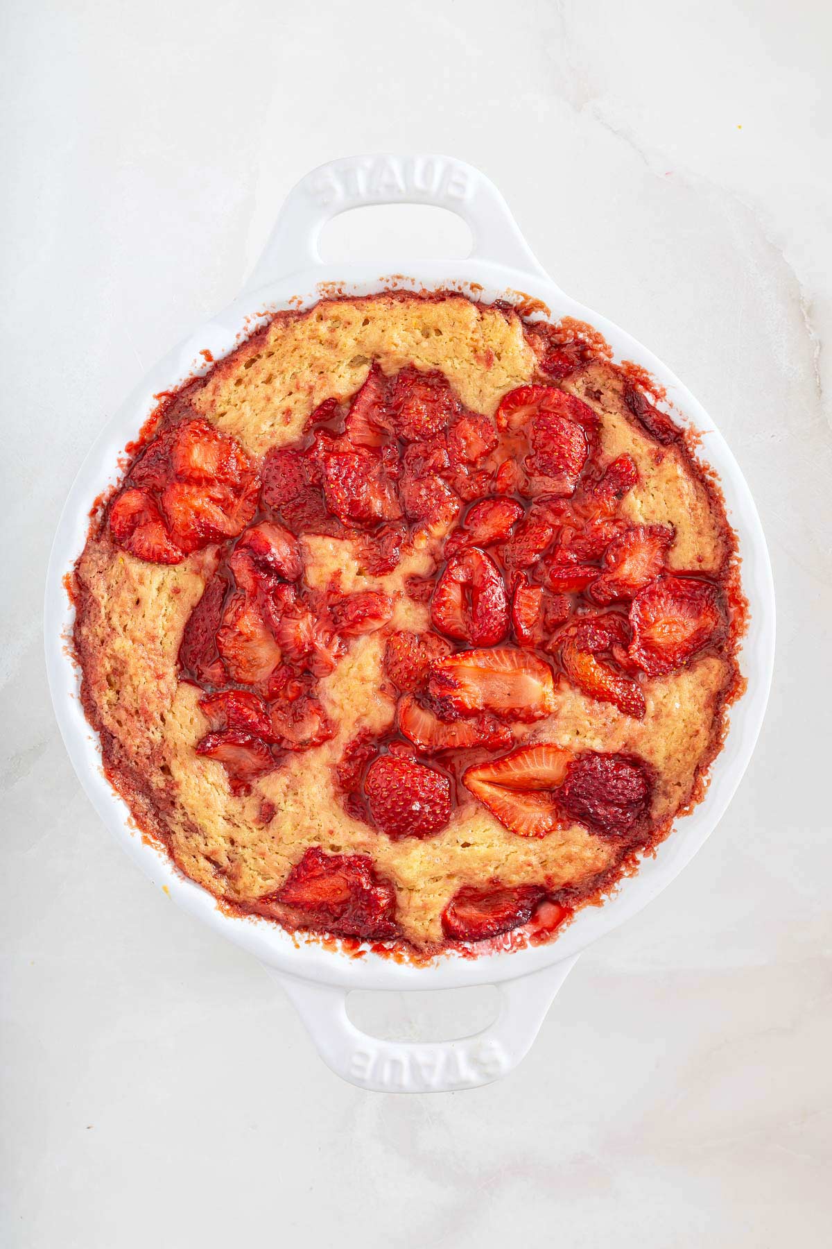 Baked strawberry oatmeal casserole in a white round dish, reminiscent of a sheetpan pancake, topped with sliced strawberries, on a light marble surface.