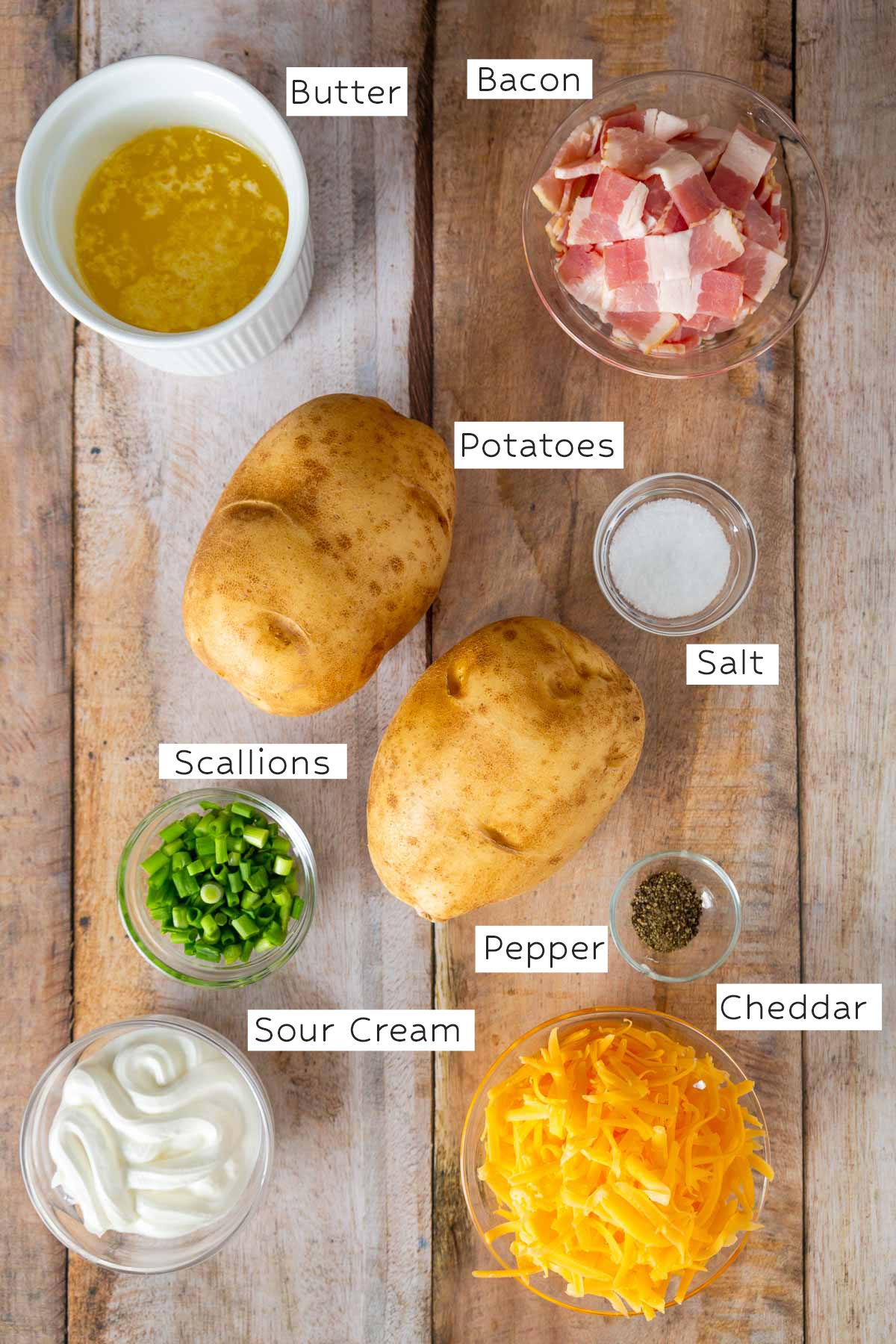 Various ingredients for baked potatoes, including butter, bacon, two potatoes, salt, pepper, scallions, sour cream, and shredded cheddar cheese, arranged on a wooden surface-perfect accompaniments for an easy sheetpan pancake recipe or hearty sheet pan pancakes.