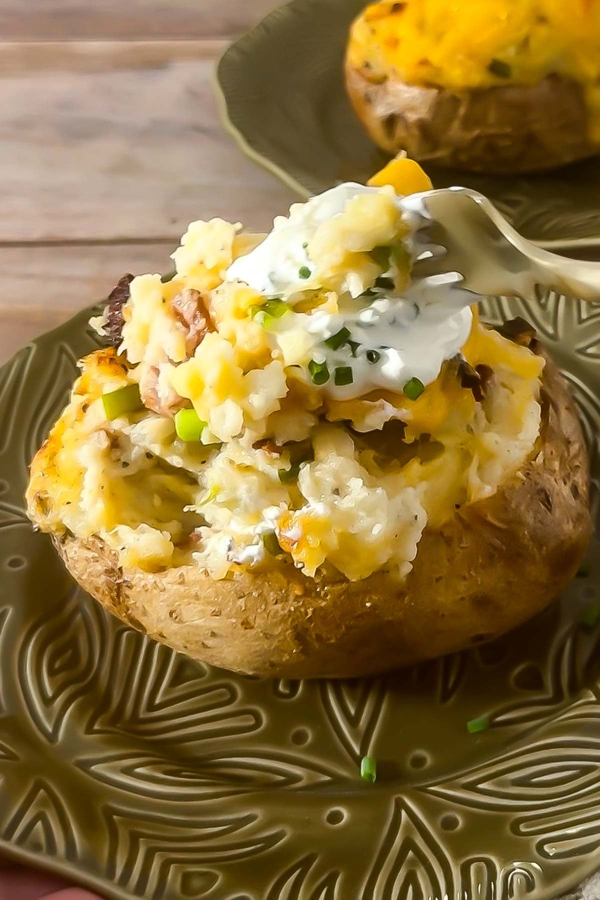 A baked potato topped with sour cream, chopped chives, cheese, and bacon sits on a green plate-echoing the hearty comfort of a classic sheet pan pancake breakfast. A fork is holding a bite of the potato.