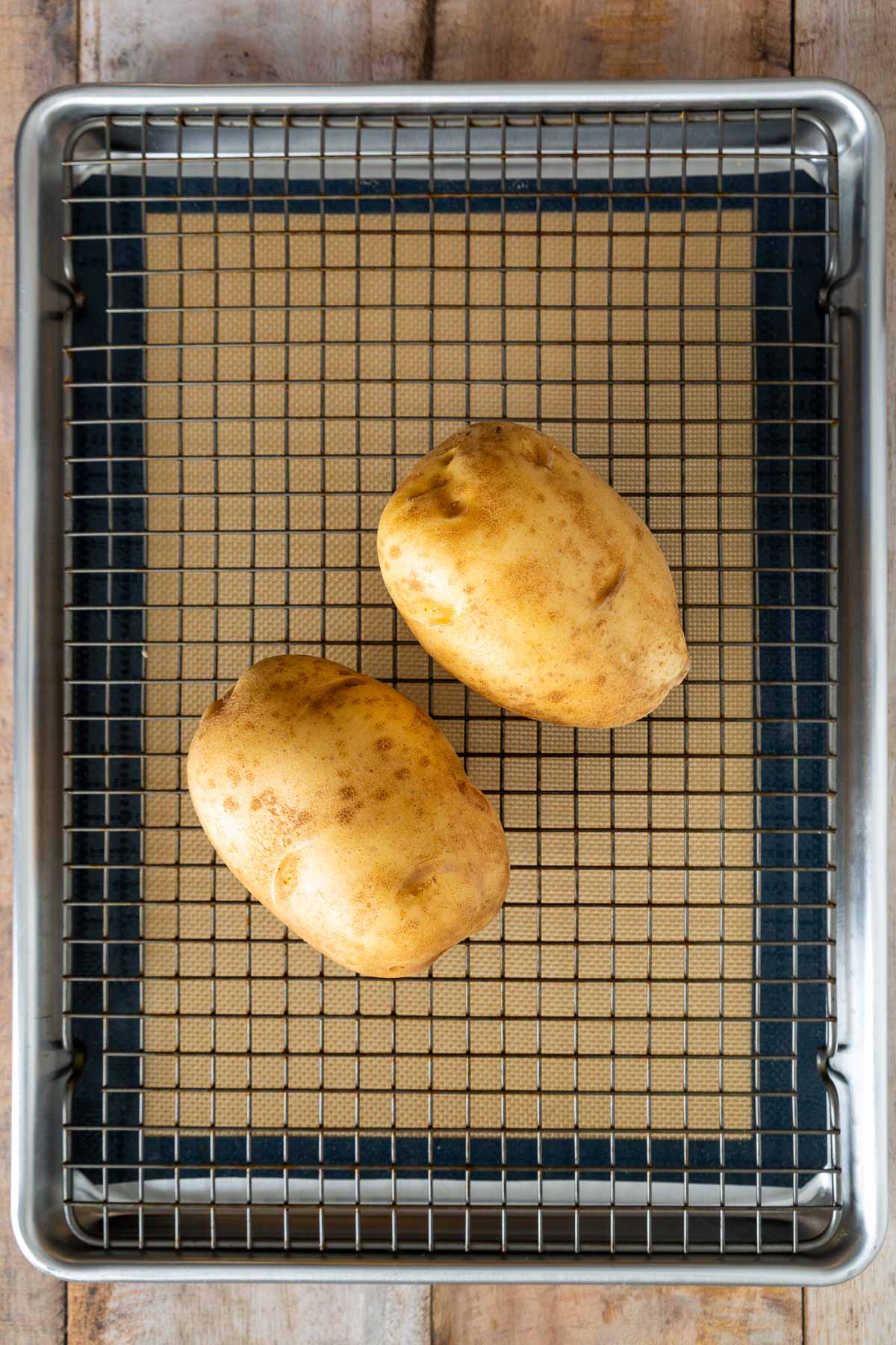 Two whole russet potatoes placed on a wire rack over a baking sheet lined with a silicone mat, reminiscent of prepping for delicious sheet pan pancakes on a wooden surface.