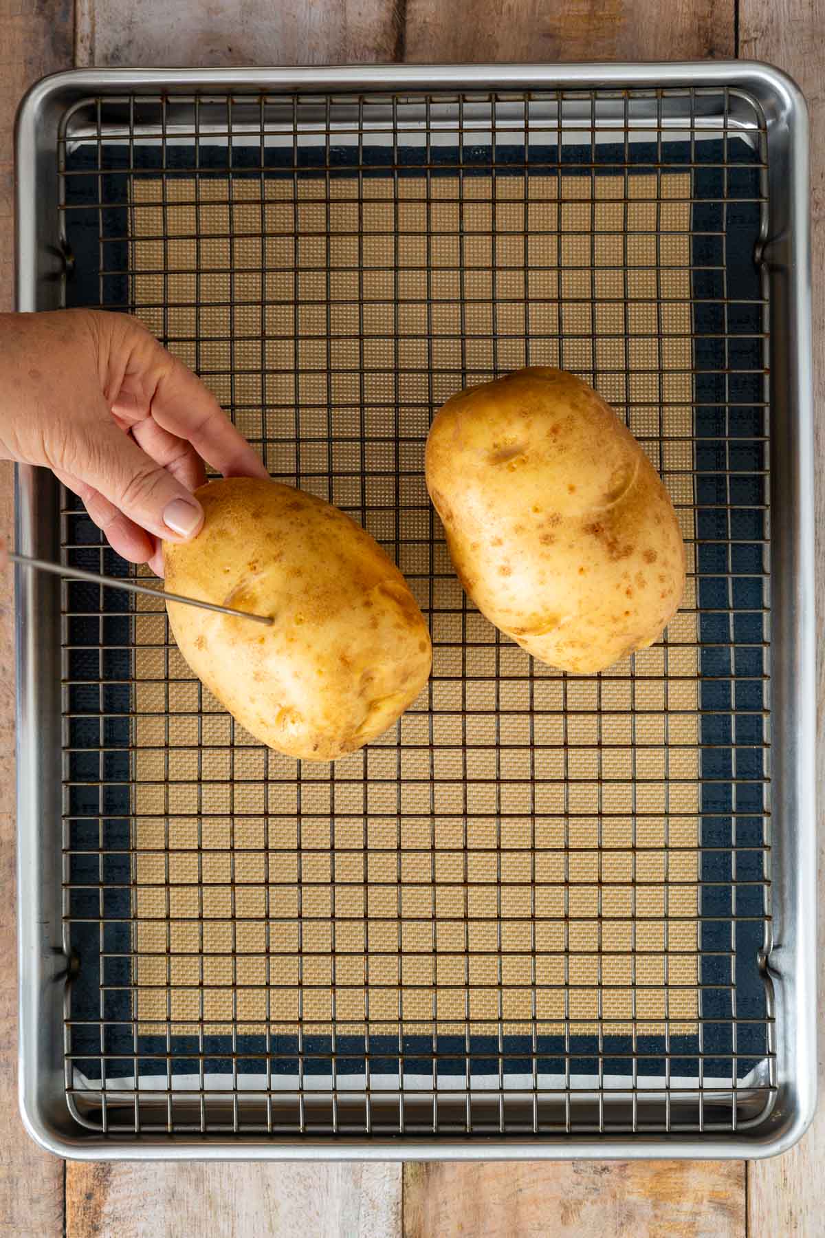 A hand pierces a raw potato with a fork on a wire rack over a baking sheet; another potato is beside it, ready to accompany dishes like fluffy sheet pan pancakes from mix.