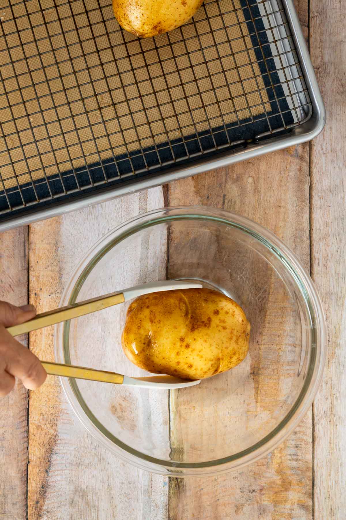 A hand uses tongs to place a whole potato into a glass bowl; a baking tray with a wire rack and mat, perfect for making sheetpan pancake recipes or preparing sheet pan pancakes from mix, is nearby on a wooden surface.