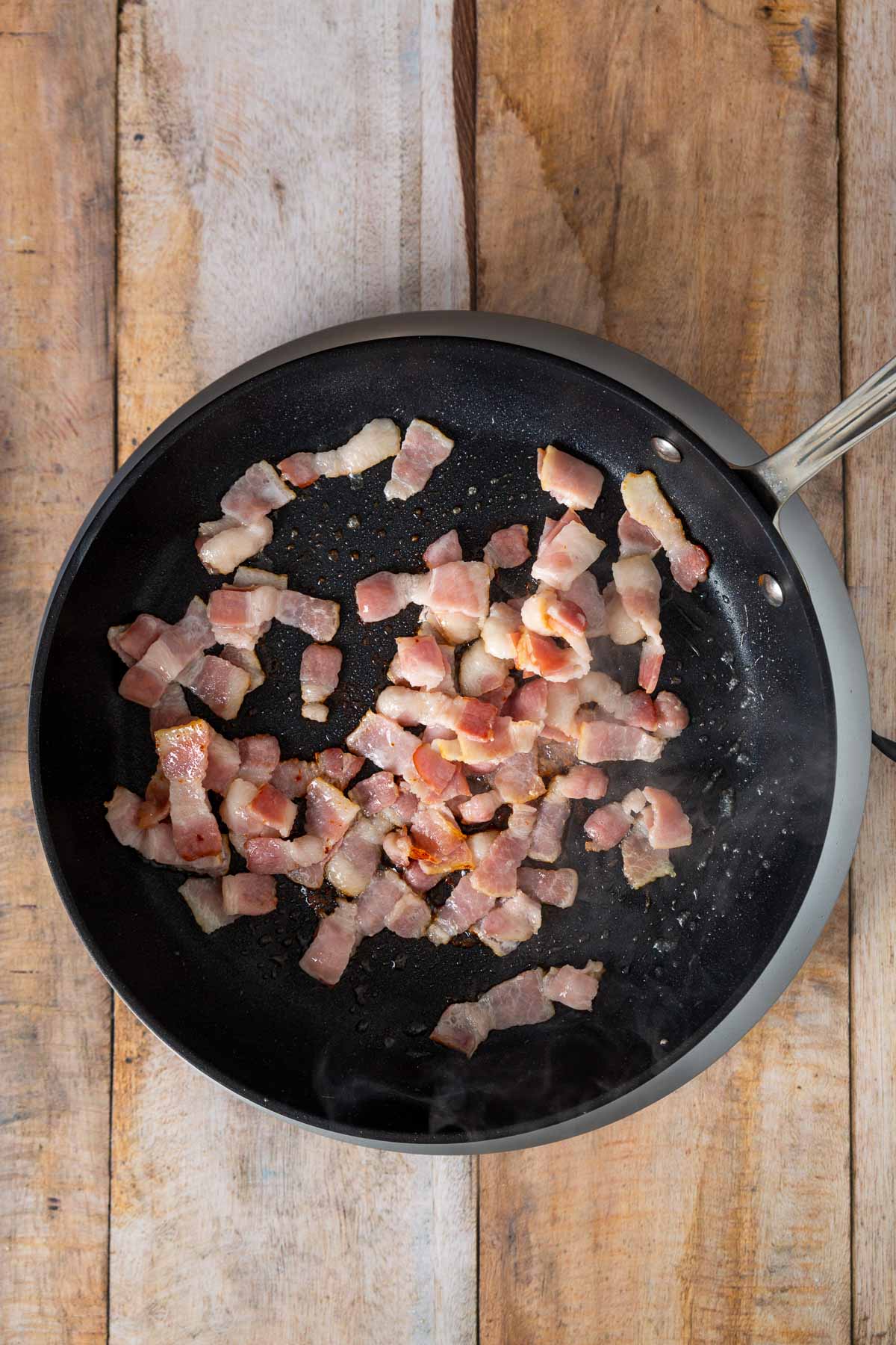 Chopped raw bacon pieces cooking in a black frying pan on a wooden surface, perfect for adding a savory twist to your easy sheetpan pancakes.