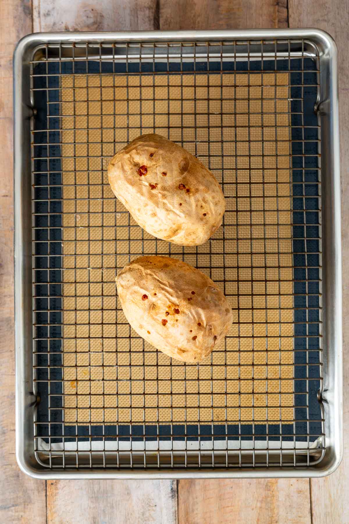 Two baked potatoes with seasoning sit on a wire rack over a silicone baking mat inside a metal baking tray on a wooden surface, ready to share the spotlight with your next sheetpan pancake recipe or easy sheet pan pancakes from mix for an effortless and delicious meal.