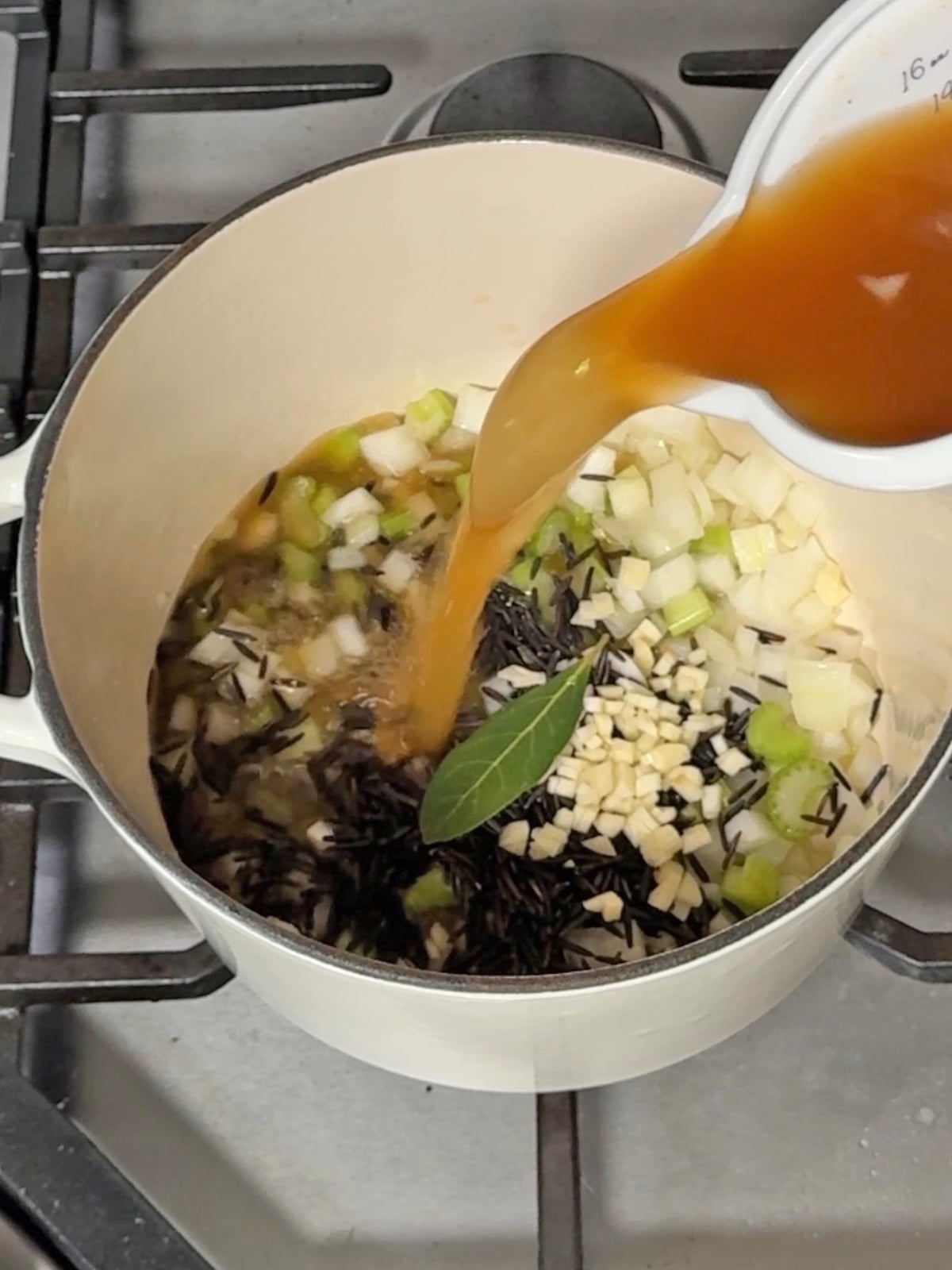 Broth being poured into a pot containing wild rice, chopped vegetables, and a bay leaf, prepared in a style as simple as easy sheet pan pancakes.